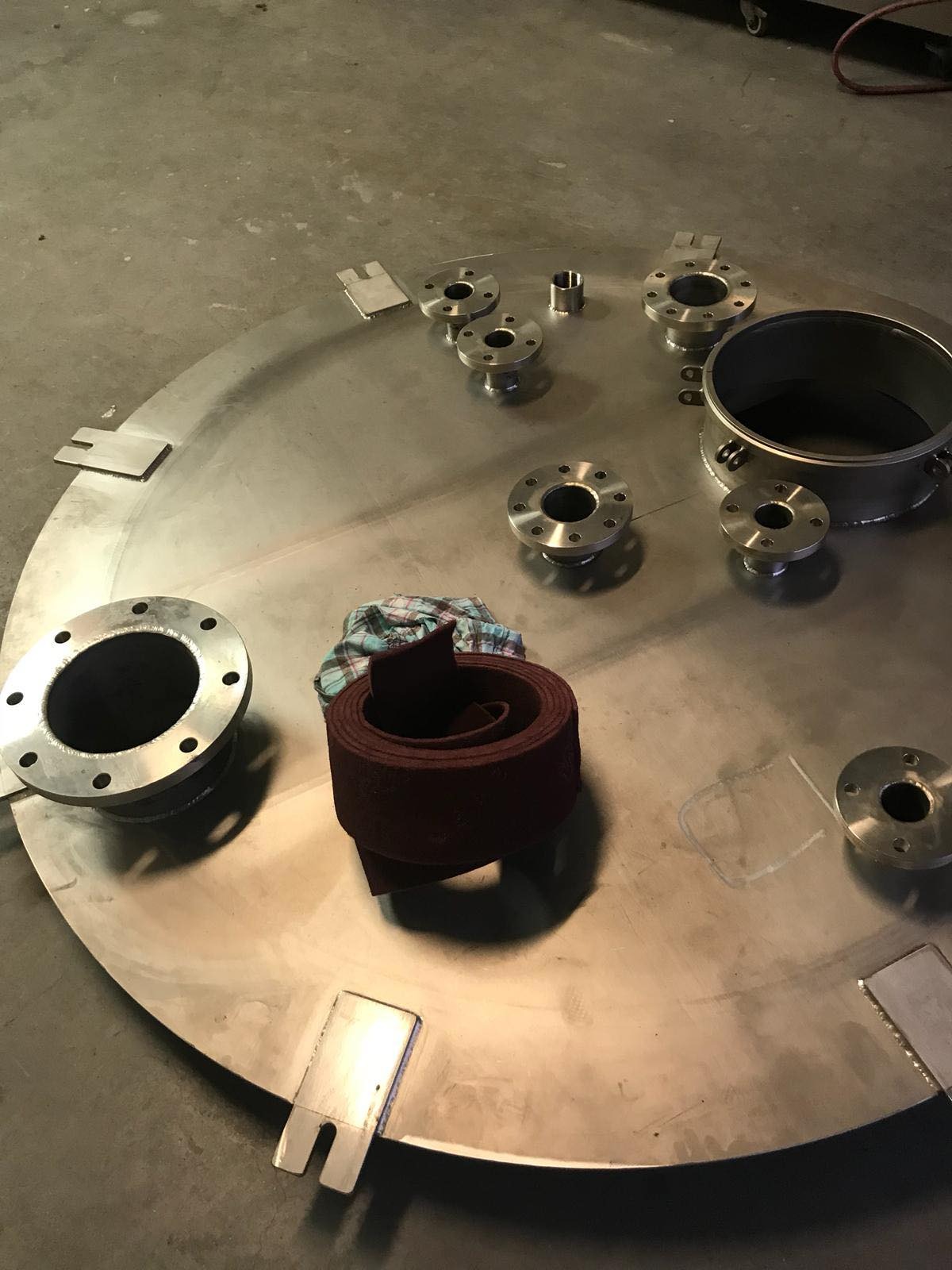 Elementy konstrukcji stalowej: okrągła płyta z wyciętymi otworami, kołnierze i zwój brązowej taśmy leżące na betonowej posadzce warsztatu.