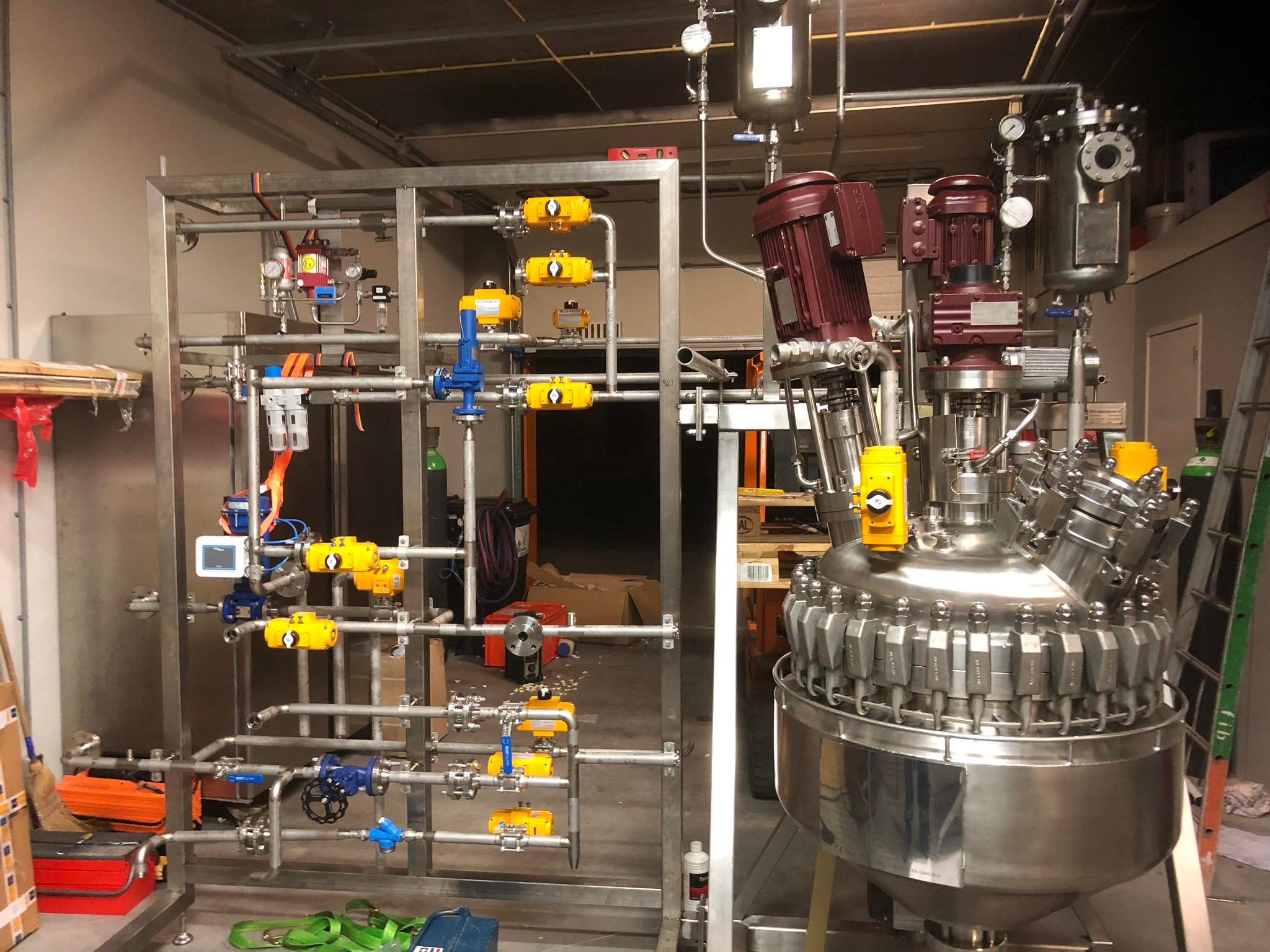 Kompleksowa instalacja przemysłowa ze stali nierdzewnej z licznymi zaworami, rurami i zbiornikiem mieszającym, widoczna w hali produkcyjnej z drabiną i skrzynkami narzędziowymi w tle.