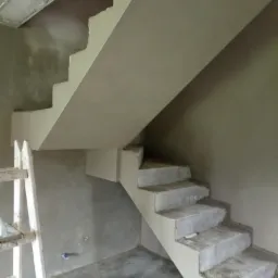 Wnętrze w trakcie prac wykończeniowych: surowe schody betonowe przygotowywane do położenia gładzi, widoczna drabina malarska i ślady zaprawy na podłodze.