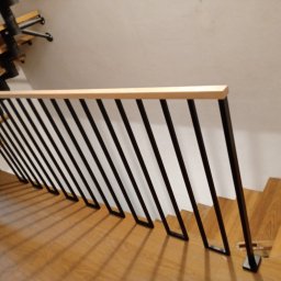 Ol-stal - Nowoczesna balustrada schodowa z czarnymi pionowymi elementami i jasnym drewnianym poręczą, zamontowana na drewnianych schodach w jasnym wnętrzu.