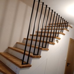 Ol-stal - Schody drewniane z czarną, nowoczesną balustradą pionową, widok z dołu, ściany pomalowane na biało, widoczna lampa na suficie i dwoje drzwi w tle.