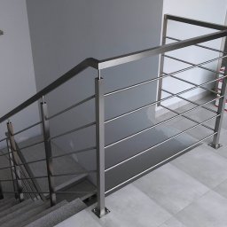 Ol-stal - Minimalistyczna balustrada nierdzewna ze stali szczotkowanej, z poziomymi prętami, zamontowana przy schodach wykończonych szarym gresem, widoczne opakowania materiałów budowlanych w tle.