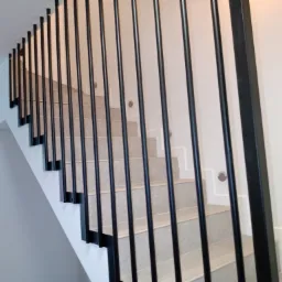 Nowoczesna balustrada schodowa z czarnych, pionowych prętów, zamocowana do stopni wyłożonych jasnymi płytkami, z oświetleniem w ścianie.