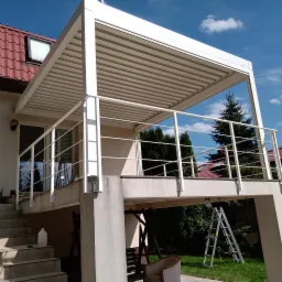 Biała pergola z zadaszeniem lamelowym i balustradą, zamontowana na betonowym tarasie przy domu z czerwoną dachówką, widok na ogród, drabina oparta o filar konstrukcji.