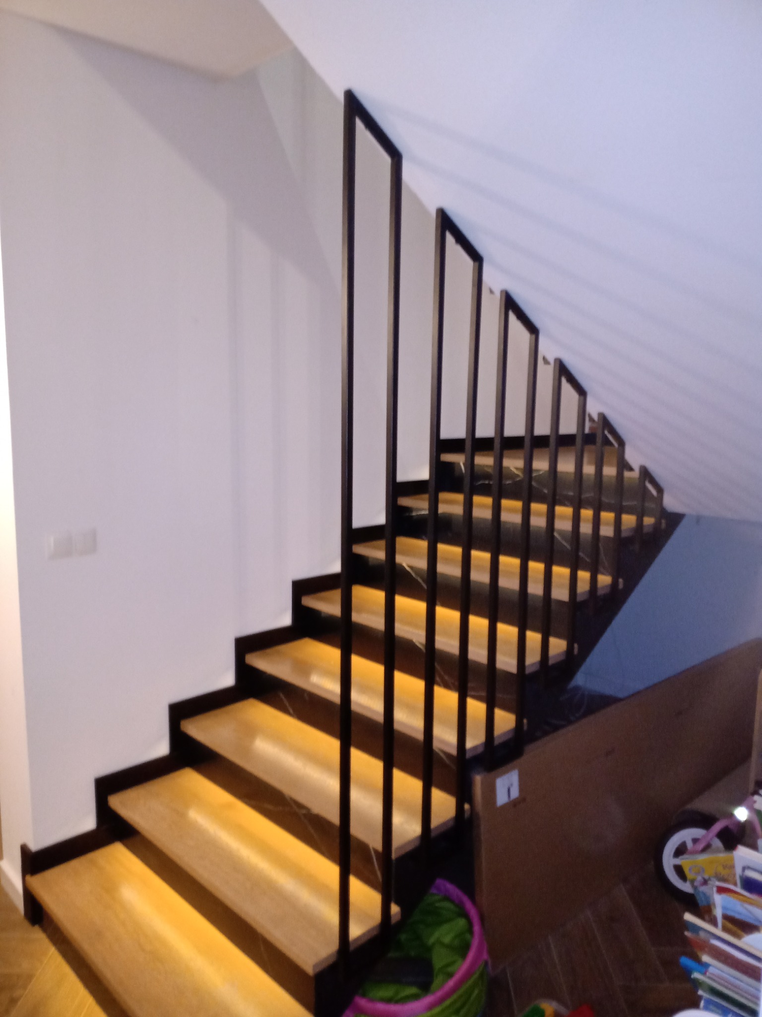 Schody wewnętrzne z drewnianymi stopniami i czarną, minimalistyczną balustradą, oświetlone od spodu, prowadzące na poddasze.