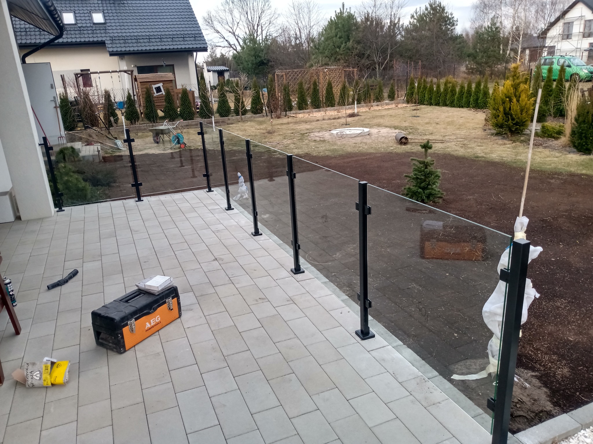 Montaż balustrady szklanej z czarnymi słupkami na tarasie z kostki brukowej, widoczna skrzynka narzędziowa AEG i elementy zabezpieczające szkło, w tle ogród i dom.