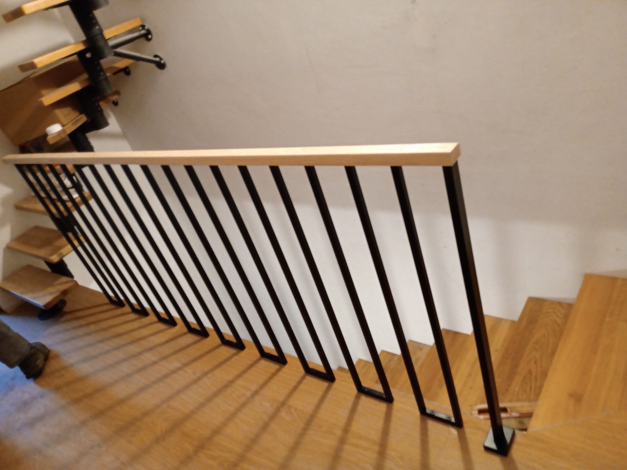 Nowoczesna balustrada schodowa z czarnymi pionowymi elementami i jasnym drewnianym poręczą, zamontowana na drewnianych schodach w jasnym wnętrzu.