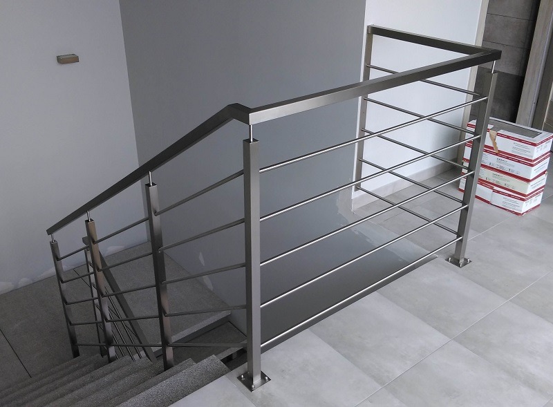 Minimalistyczna balustrada nierdzewna ze stali szczotkowanej, z poziomymi prętami, zamontowana przy schodach wykończonych szarym gresem, widoczne opakowania materiałów budowlanych w tle.