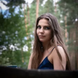 Portret młodej kobiety z długimi brązowymi włosami w lesie, ubrana w granatową sukienkę na ramiączkach, z delikatnym makijażem i intensywnym spojrzeniem.