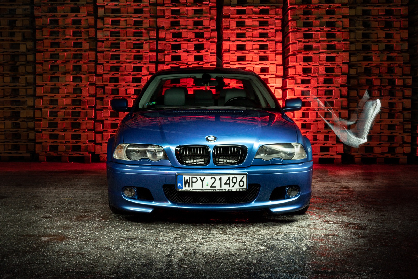 Niebieskie BMW E46 sfotografowane z przodu na tle ściany z palet oświetlonych czerwonym światłem, z efektem rozmycia ruchu po prawej stronie.