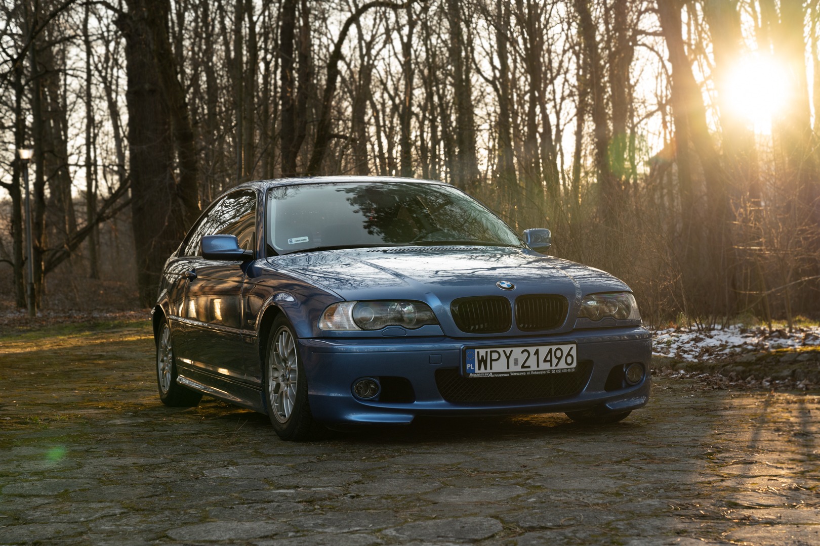 Niebieskie BMW E46 na tle drzew w słoneczny dzień, widoczne tablice rejestracyjne WPY 21496 z adresem warsztatu samochodowego.