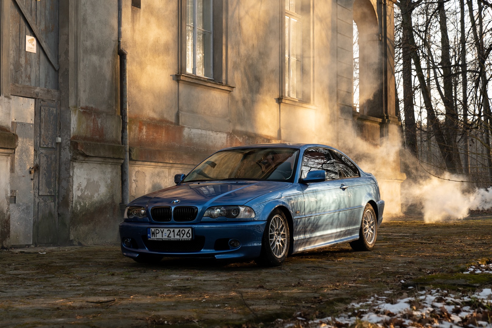 Błękitny samochód BMW E46 Coupe, zaparkowany przed starą kamienicą z drewnianymi drzwiami i oknami, otoczony mgłą i drzewami bez liści.