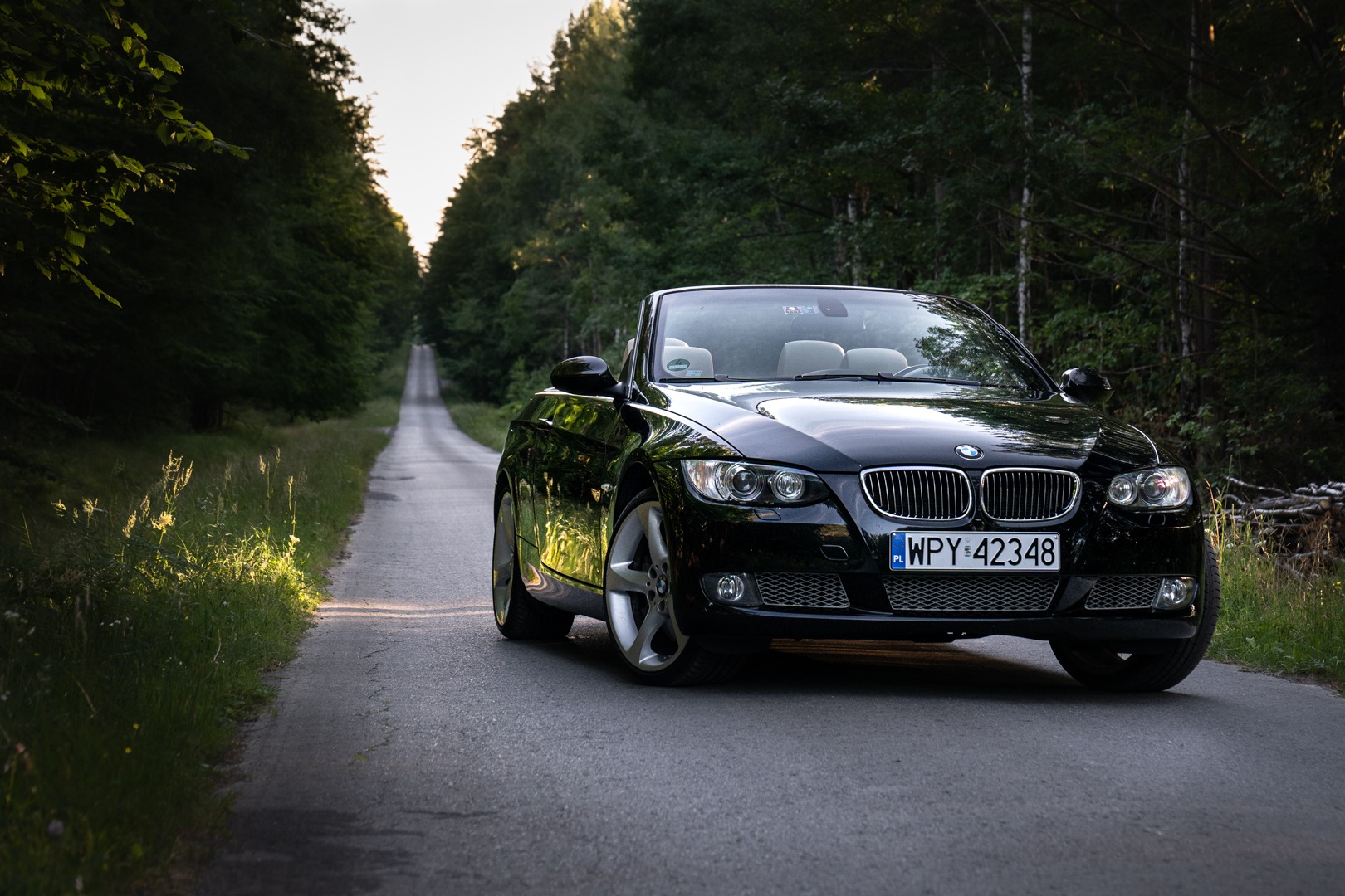 Czarny kabriolet BMW z beżowym wnętrzem, zaparkowany na wąskiej asfaltowej drodze prowadzącej przez gęsty las, tablica rejestracyjna WPY 42348.