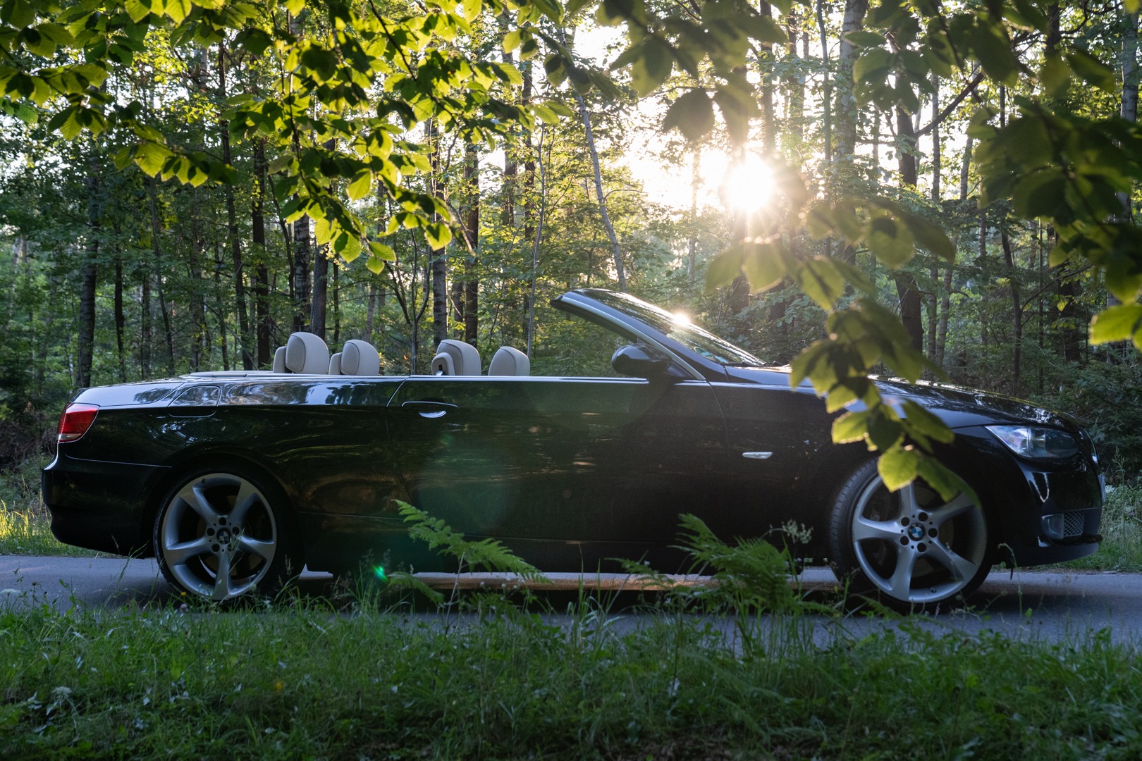 Czarny kabriolet z jasnymi siedzeniami, sfotografowany w Radomiu na tle lasu w słoneczny dzień, z liśćmi w pierwszym planie.
