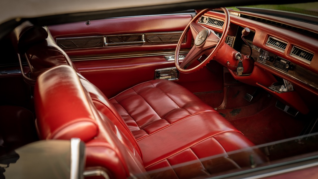 Wnętrze klasycznego, czerwonego kabrioletu Cadillac z widocznym skórzanym siedzeniem, kierownicą z logo i deską rozdzielczą z elementami drewna.