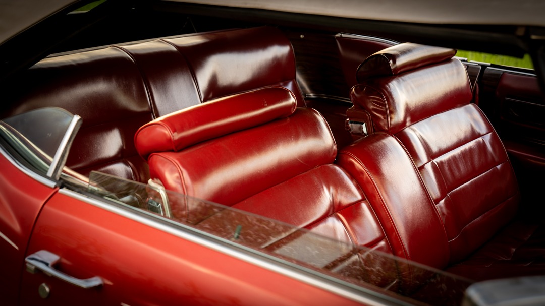 Wnętrze klasycznego, czerwonego kabrioletu z lśniącymi, skórzanymi siedzeniami, widoczne przez lekko zabrudzoną szybę boczną.