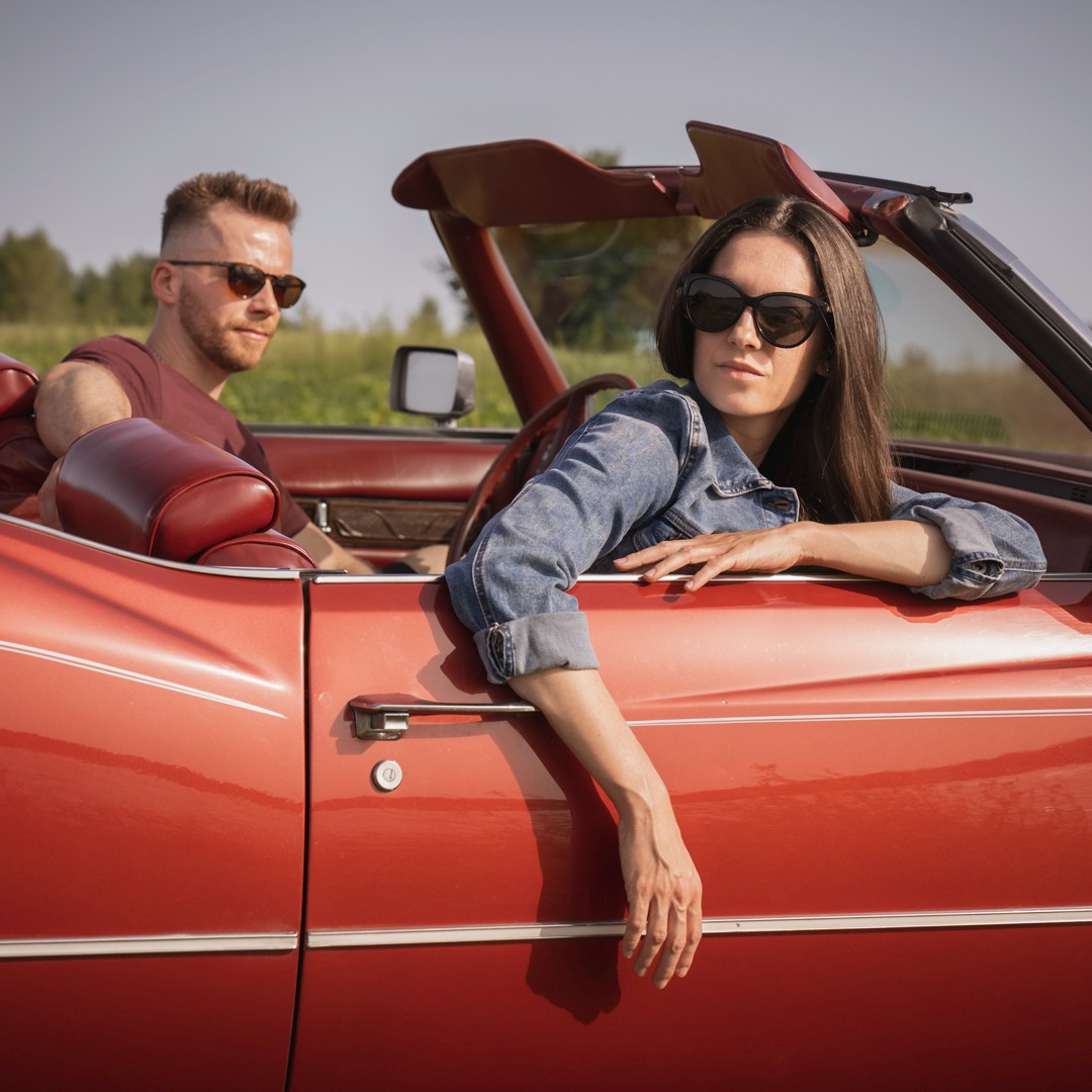 Para w okularach przeciwsłonecznych podróżuje czerwonym kabrioletem w słoneczny dzień, kobieta opiera rękę o drzwi.