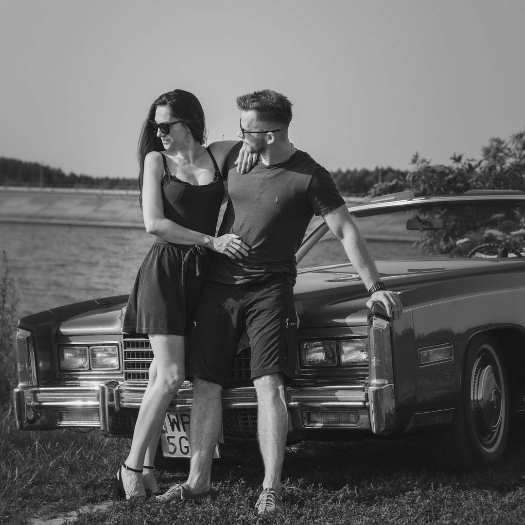 Czarno-białe zdjęcie pary w okularach przeciwsłonecznych, pozującej przy klasycznym samochodzie na tle zbiornika wodnego.