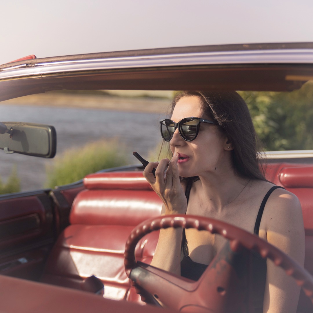 Kobieta w okularach przeciwsłonecznych maluje usta szminką w czerwonym kabriolecie z widokiem na rzekę.