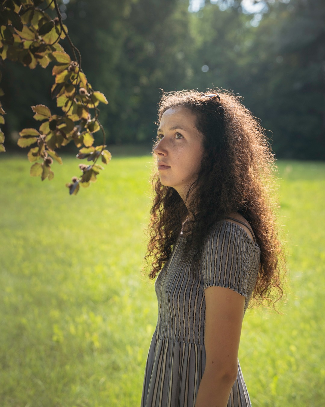 Portret kobiety z kręconymi włosami w letniej sukience, stojącej na tle zielonej trawy i drzew, z gałęzią z liśćmi w lewym górnym rogu kadru.