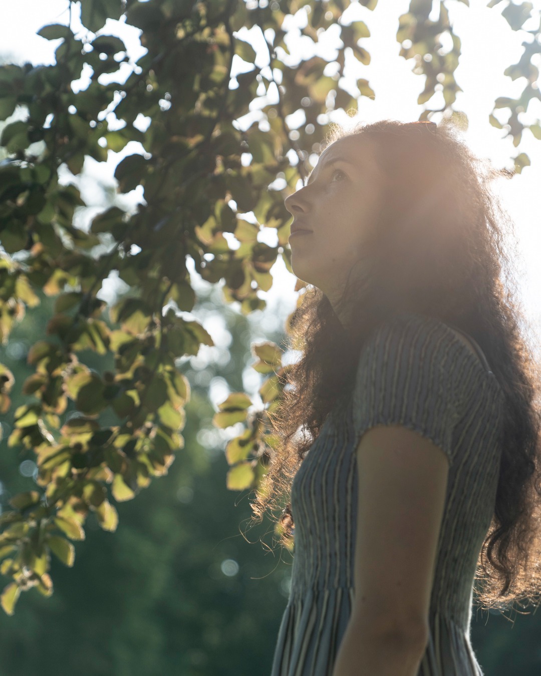 Portret kobiety z kręconymi włosami, spoglądającej w górę, w promieniach słońca przebijających przez liście drzewa.