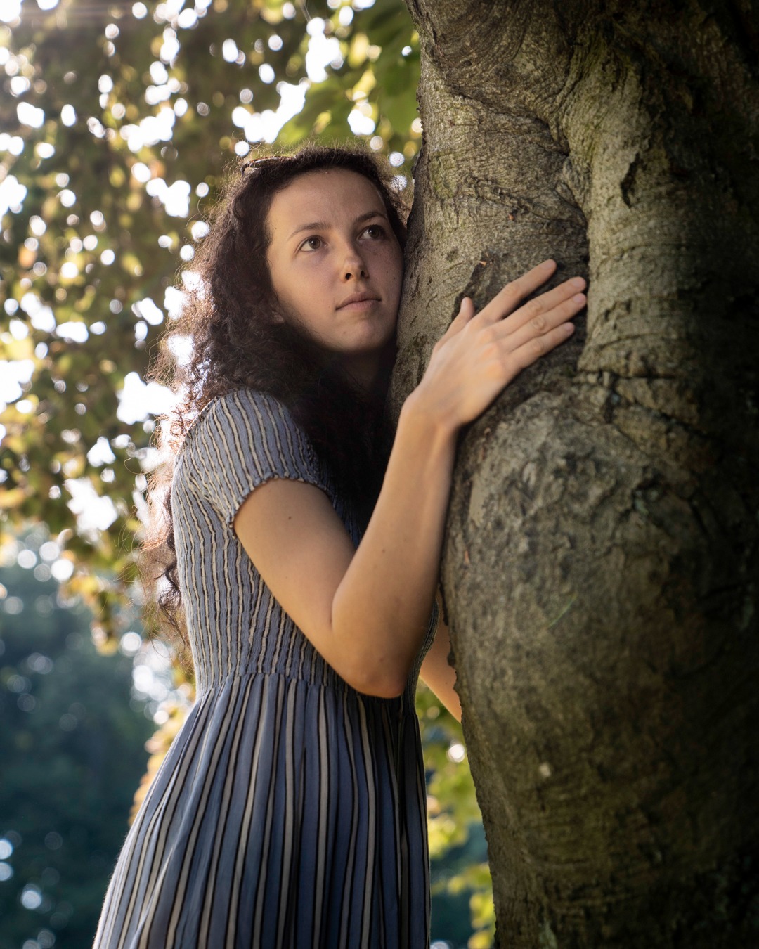 Portret kobiety w paski sukience, opierającej się o korę drzewa, spoglądającej w górę w letnim świetle.