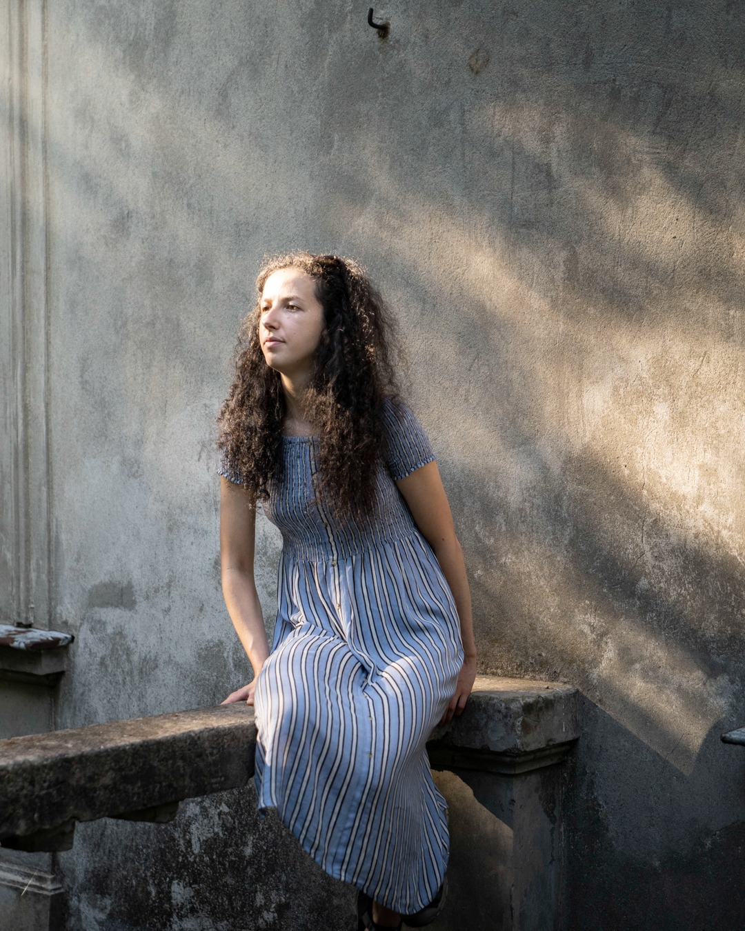 Portret kobiety w niebieskiej sukience w paski, siedzącej na kamiennym murku, z promieniem słońca padającym na tło ściany.