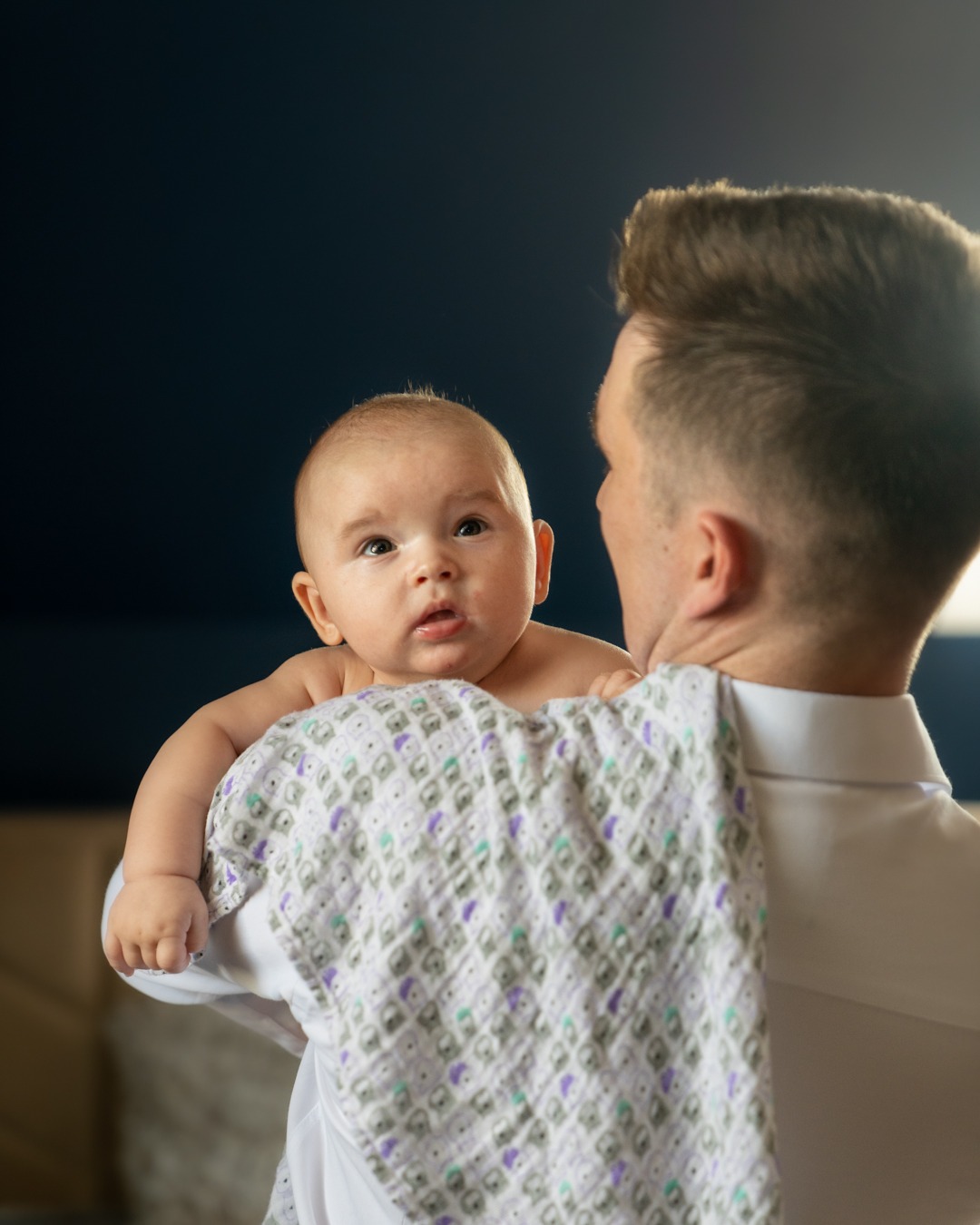 Ojciec w białej koszuli trzyma na ramieniu niemowlę owinięte w pieluszkę w geometryczne wzory, dziecko patrzy prosto w obiektyw.