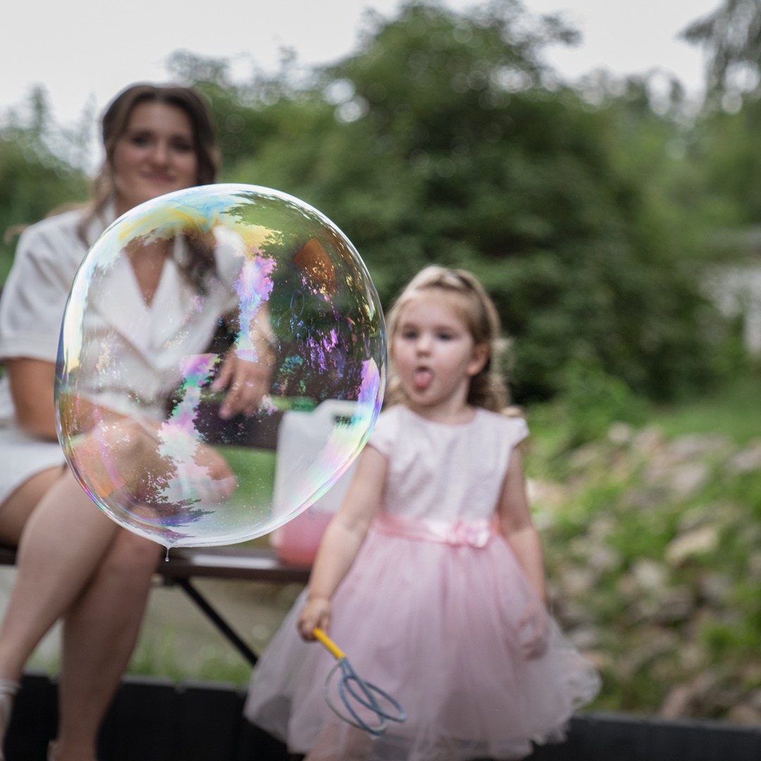 Dziewczynka w różowej sukience puszcza ogromne bańki mydlane, w tle siedząca kobieta na ławce i zieleń.