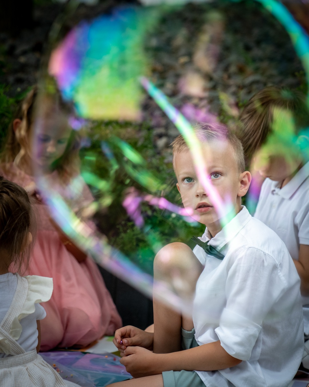 Chłopiec w białej koszuli i muszce siedzi na kocu z innymi dziećmi, widoczny przez dużą, tęczową bańkę mydlaną.
