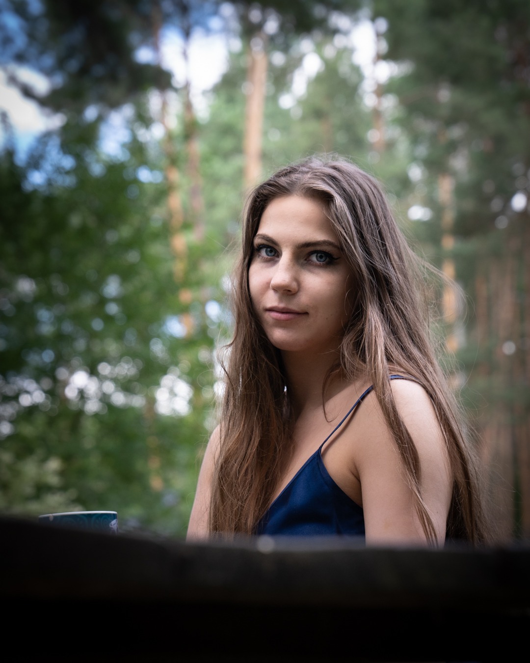 Portret młodej kobiety z długimi brązowymi włosami w lesie, ubrana w granatową sukienkę na ramiączkach, z delikatnym makijażem i intensywnym spojrzeniem.