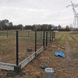 Montaż Bram i ogrodzeń Arendt Rafał - Częściowo zamontowane czarne ogrodzenie z siatki z betonową podmurówką na działce, widoczne narzędzia i kanister, w tle słup wysokiego napięcia i domy.