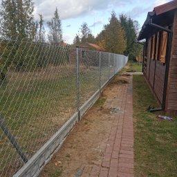 Montaż Bram i ogrodzeń Arendt Rafał - Długa siatka ogrodzeniowa z podmurówką betonową, rozciągająca się wzdłuż ścieżki z czerwonej kostki brukowej i drewnianego domku w tle, widok z boku.