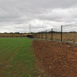 Montaż Bram i ogrodzeń Arendt Rafał - Nowo postawione, czarne ogrodzenie z paneli na betonowej podmurówce oddziela świeżo posianą trawę od brązowej ziemi uprawnej pod pochmurnym niebem.