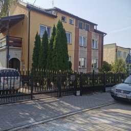 Montaż Bram i ogrodzeń Arendt Rafał - Czarna metalowa brama wjazdowa z dekoracyjnymi złotymi elementami, prowadząca do posesji z budynkami w tle, z zaparkowanymi samochodami po obu stronach.