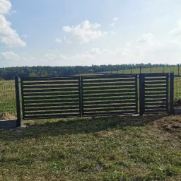 Nowoczesna, pozioma, grafitowa brama wjazdowa z furtką, widok z zewnątrz na tle łąki i lasu, brama zamontowana na betonowej podmurówce, dodatkowo widoczna siatka ogrodzeniowa.