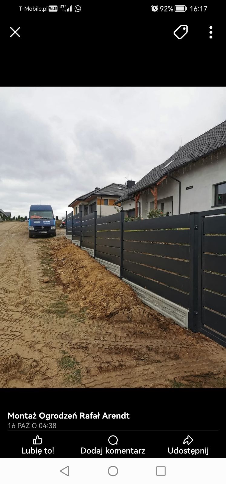 Nowoczesne, poziome przęsła w kolorze grafitowym, z podmurówką imitującą kamień, odgradzające posesję od błotnistej drogi, na tle zabudowy jednorodzinnej i firmowego busa.