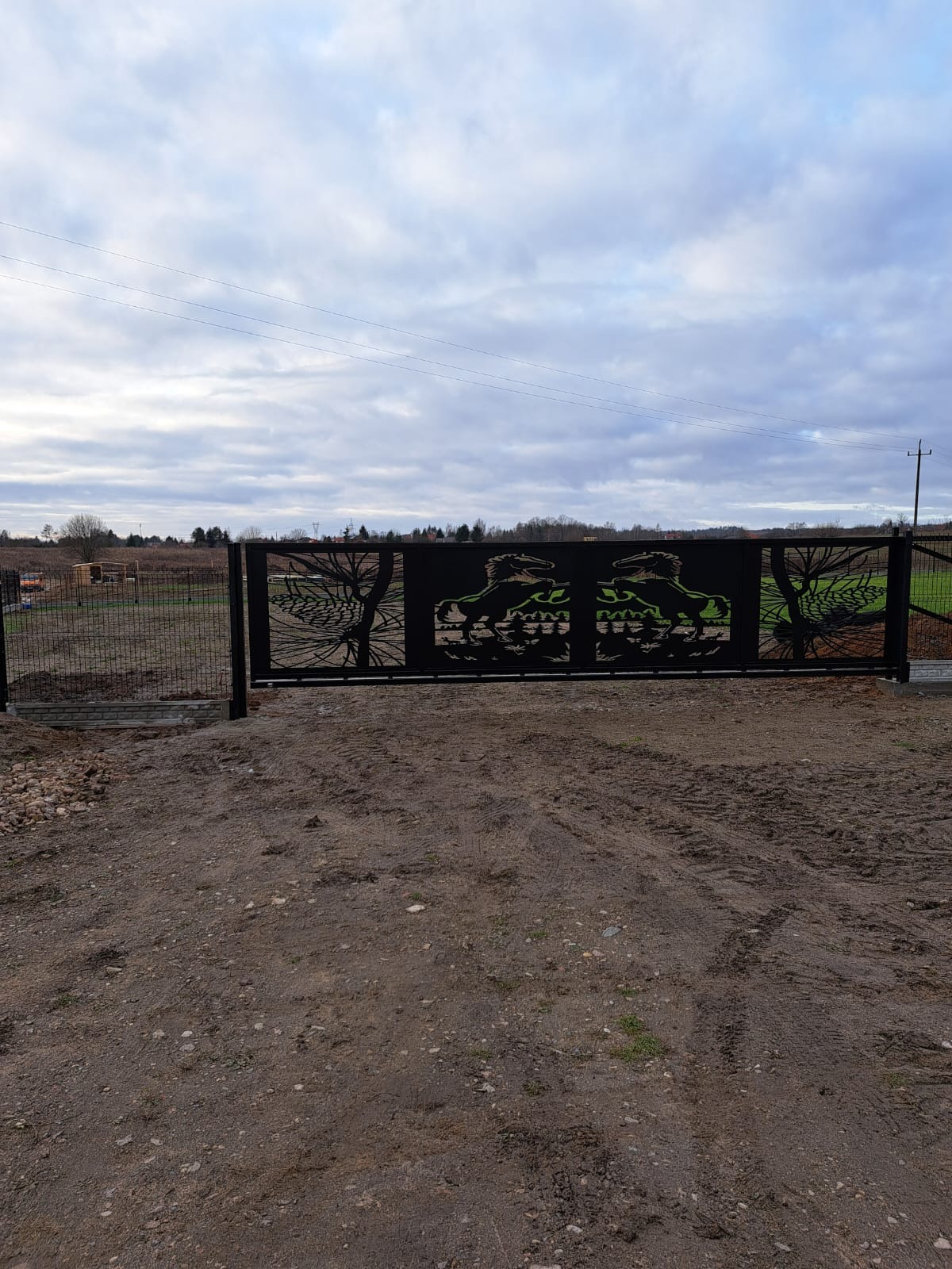 Czarna brama wjazdowa z ażurowym wzorem koni i drzew, widoczna na tle pochmurnego nieba i pola.