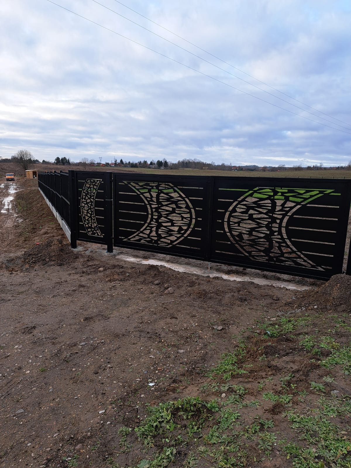 Długi, czarny system ogrodzeniowy z dekoracyjnymi, ażurowymi wzorami w kształcie półksiężyców, widoczny na tle pochmurnego nieba i polnego krajobrazu.