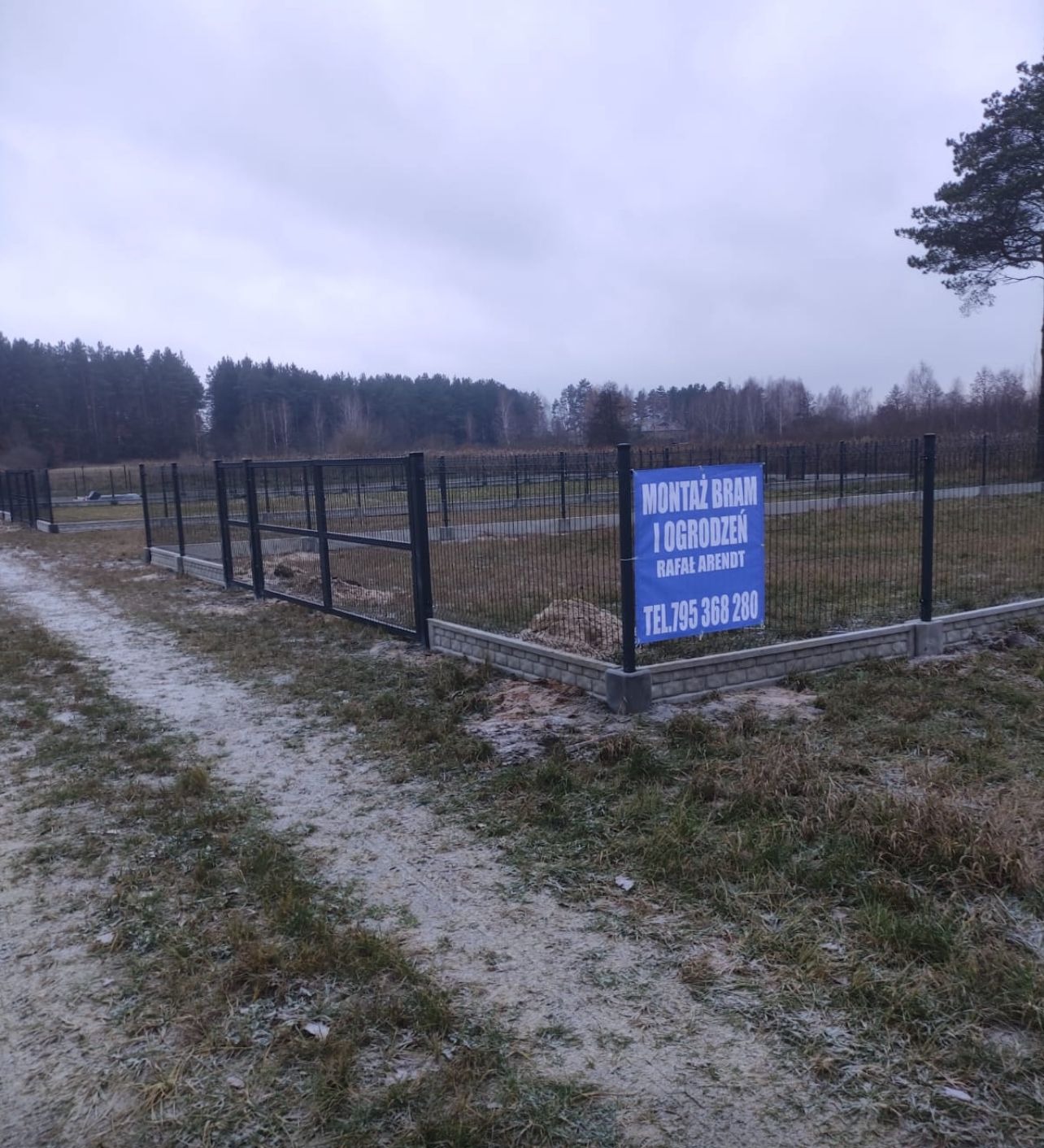 Nowo postawione czarne panele ogrodzeniowe na betonowej podmurówce, widoczne na tle zalesionego terenu w pochmurny dzień; na pierwszym planie tablica informacyjna z napisem 'Montaż bram i ogrodzeń'...