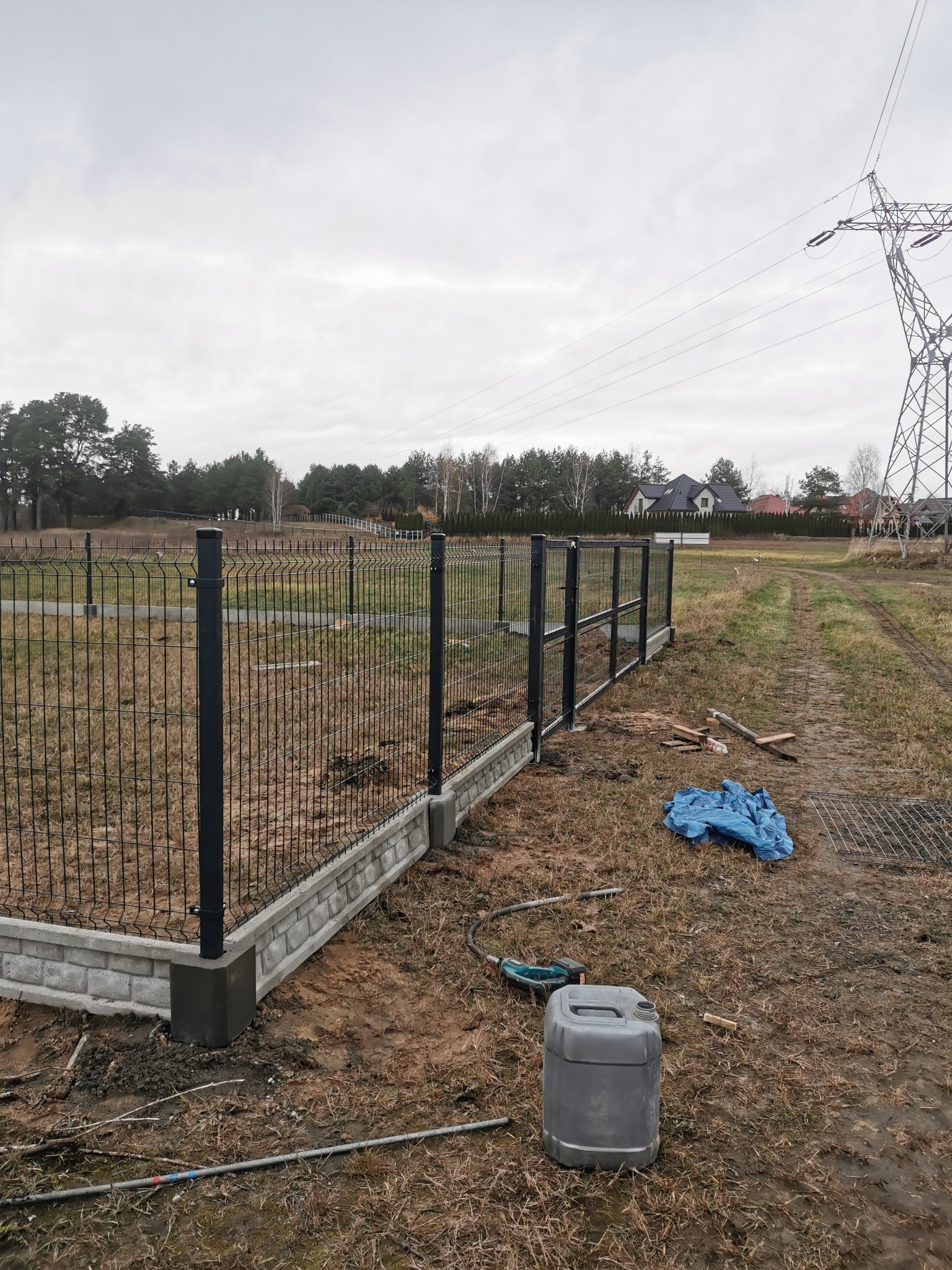 Częściowo zamontowane czarne ogrodzenie z siatki z betonową podmurówką na działce, widoczne narzędzia i kanister, w tle słup wysokiego napięcia i domy.