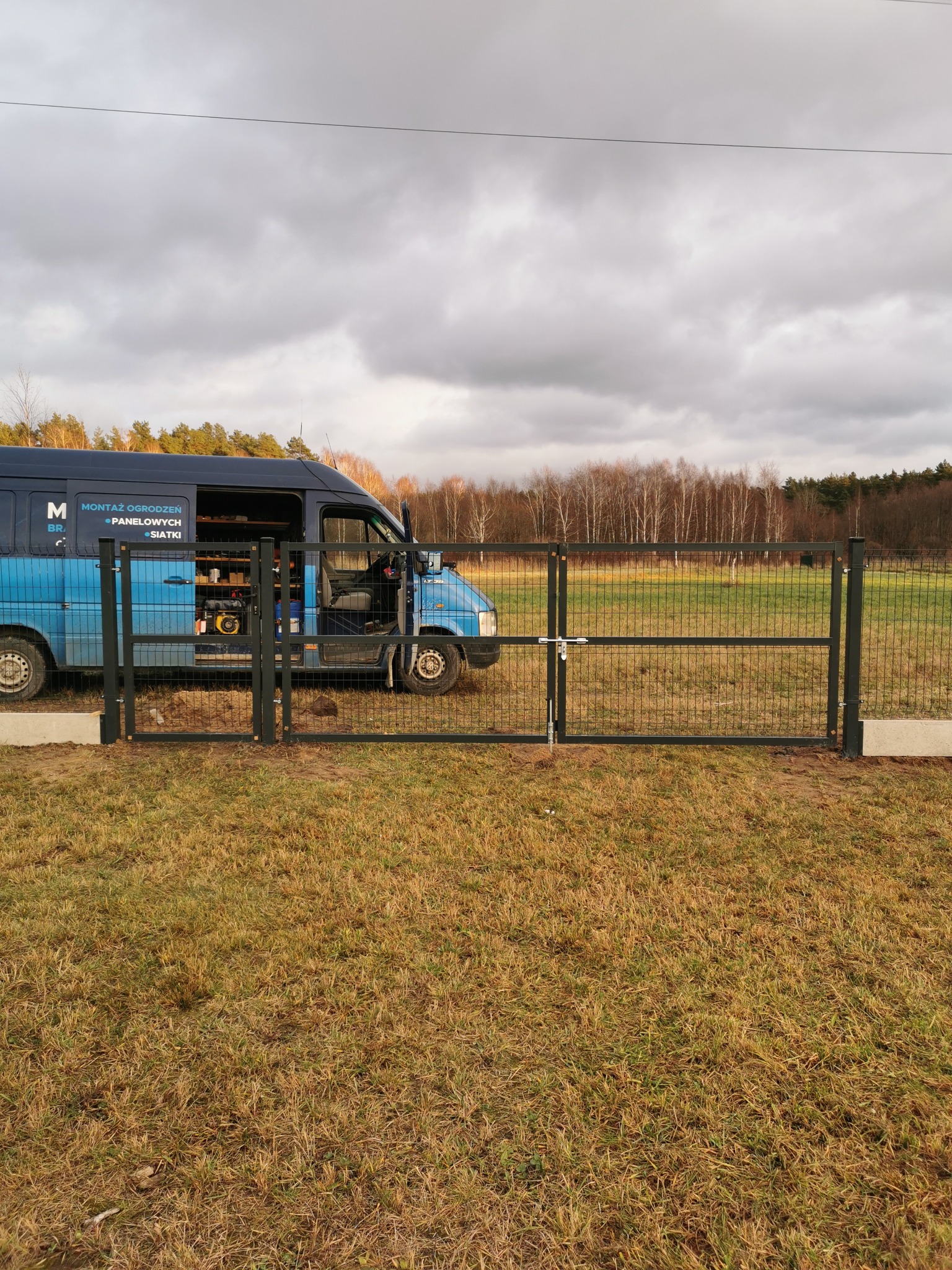 Zamknięta brama panelowa z metalową siatką i prostym zamkiem, na tle łąki i zaparkowanego firmowego busa z otwartymi drzwiami, widok z poziomu gruntu, pochmurne niebo.
