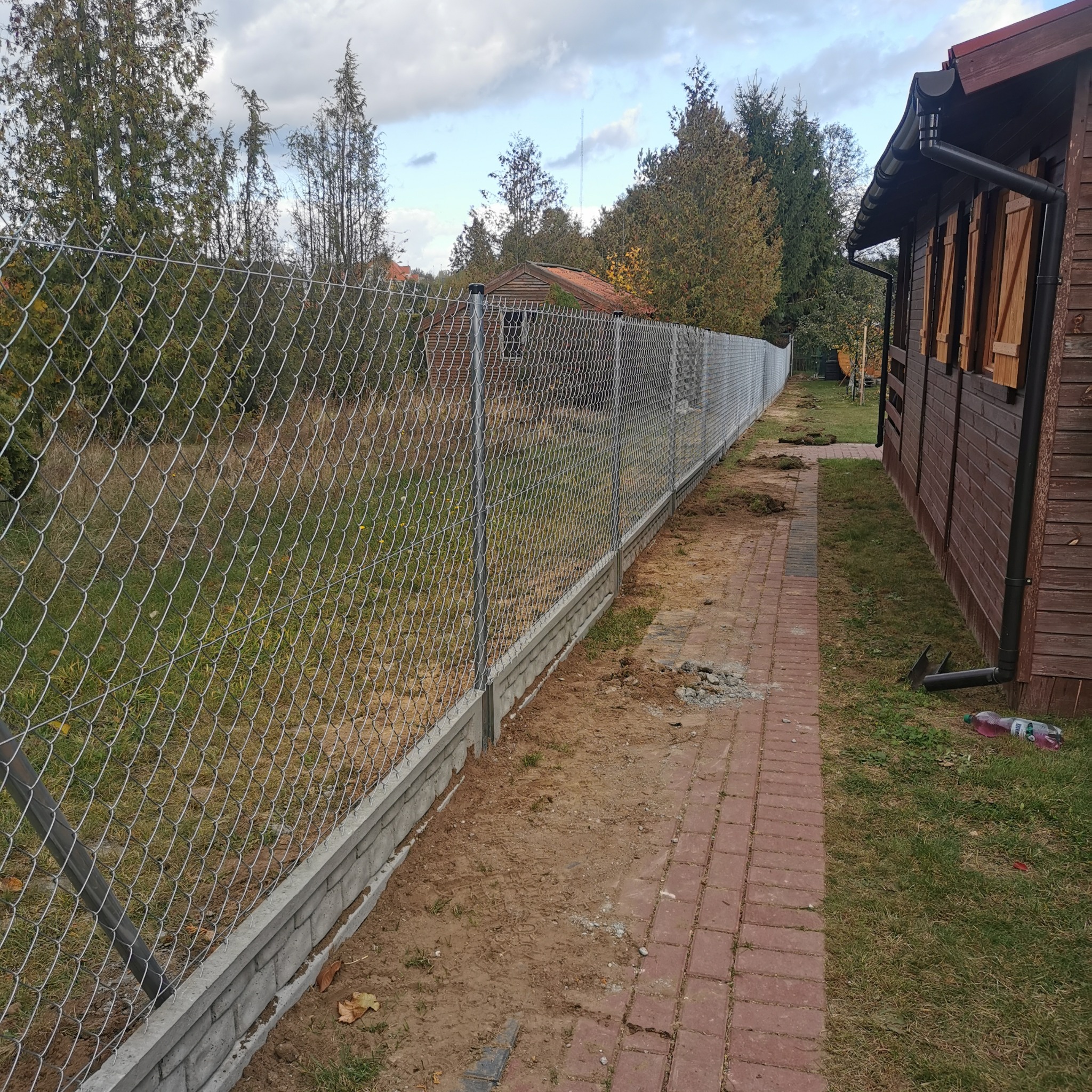 Długa siatka ogrodzeniowa z podmurówką betonową, rozciągająca się wzdłuż ścieżki z czerwonej kostki brukowej i drewnianego domku w tle, widok z boku.