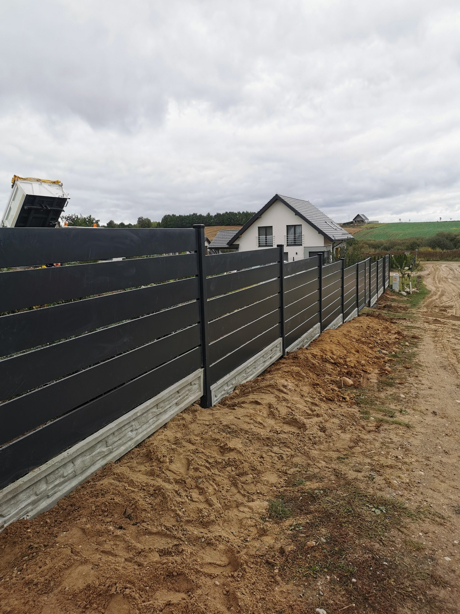 Nowoczesne, czarne ogrodzenie z poziomych desek na betonowej podmurówce, widok z boku, w tle dom jednorodzinny i wywrotka z piaskiem.