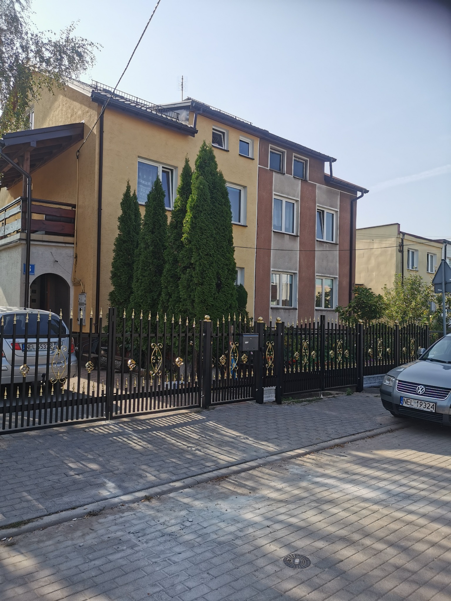 Czarna metalowa brama wjazdowa z dekoracyjnymi złotymi elementami, prowadząca do posesji z budynkami w tle, z zaparkowanymi samochodami po obu stronach.