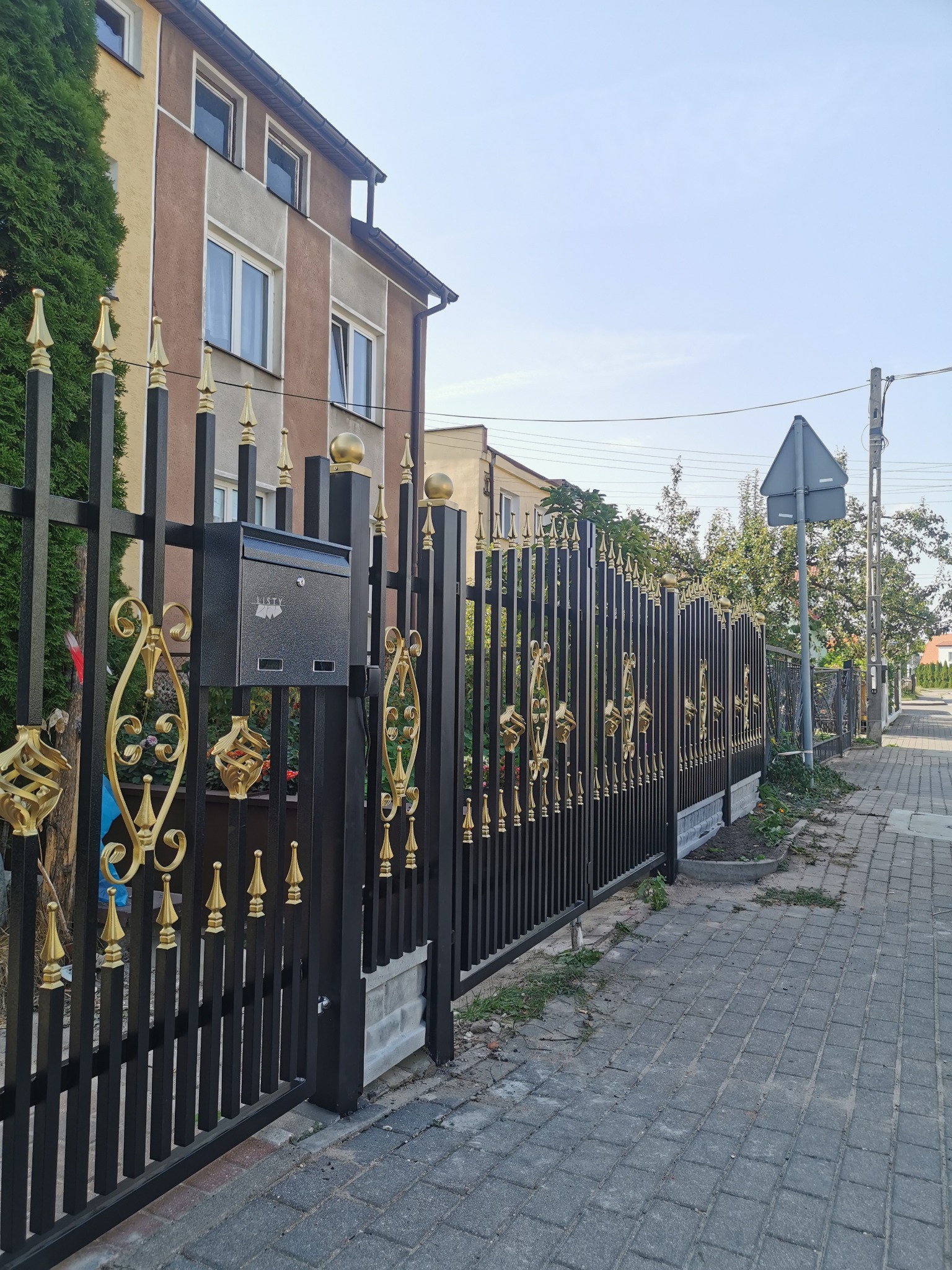Eleganckie, czarne ogrodzenie metalowe z ozdobnymi, złotymi elementami i skrzynką na listy, widok z chodnika.