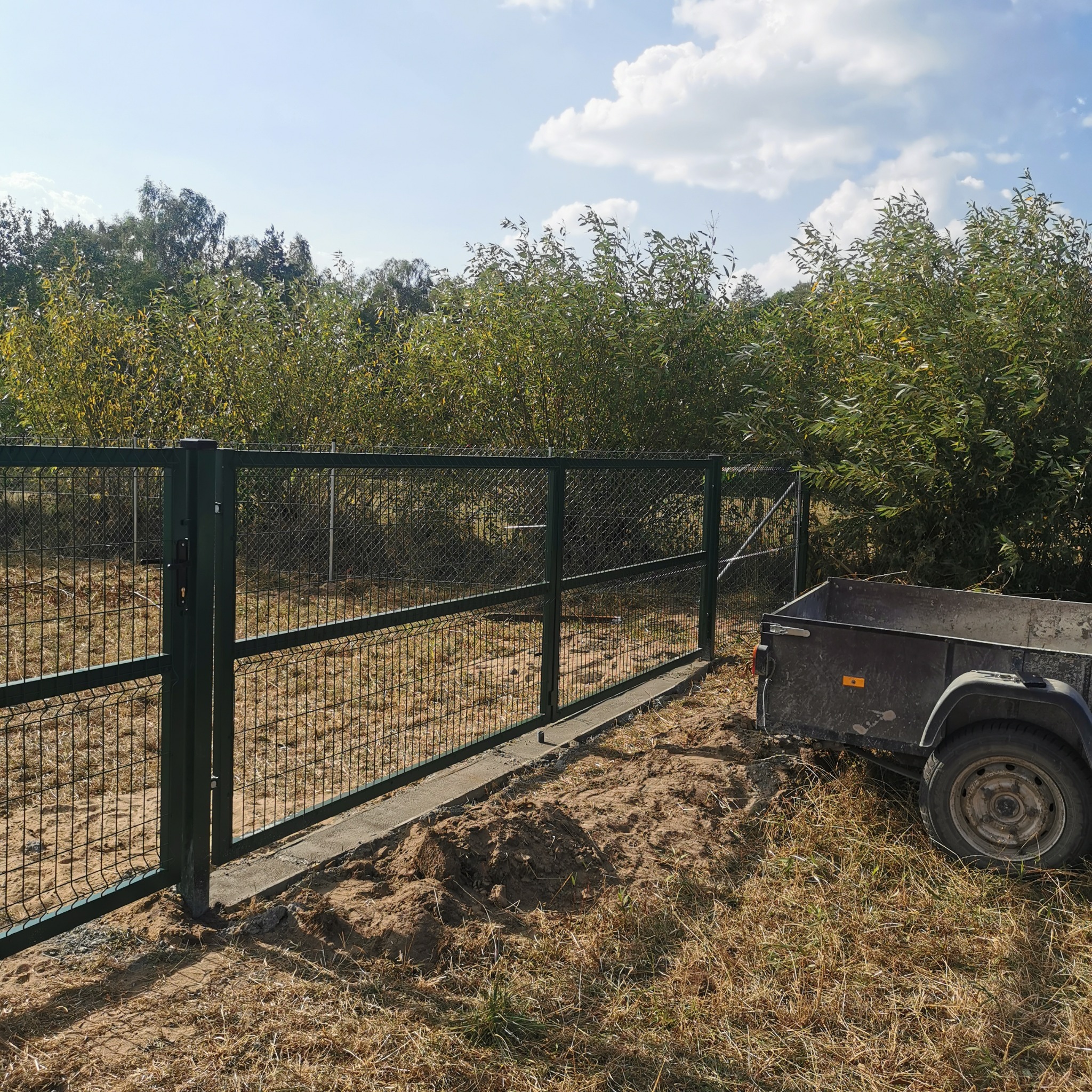 Zielona brama z siatki panelowej i przyczepka na tle drzew i nieba z chmurami, widoczne ślady prac ziemnych.
