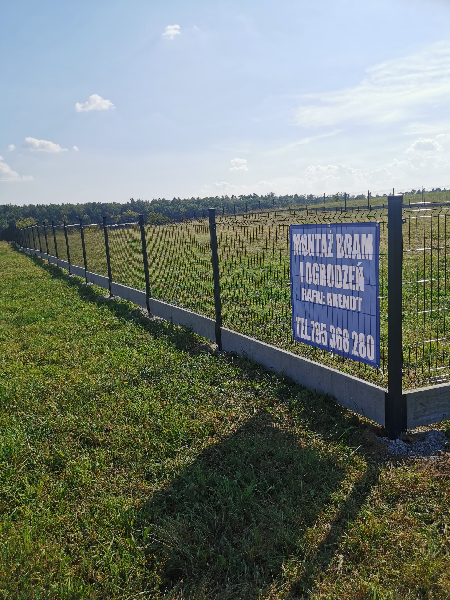 Długi, czarny panelowy płot na betonowej podmurówce, z niebieską tabliczką reklamową firmy 'Montaż Bram i Ogrodzeń Rafał Arendt' z numerem telefonu.