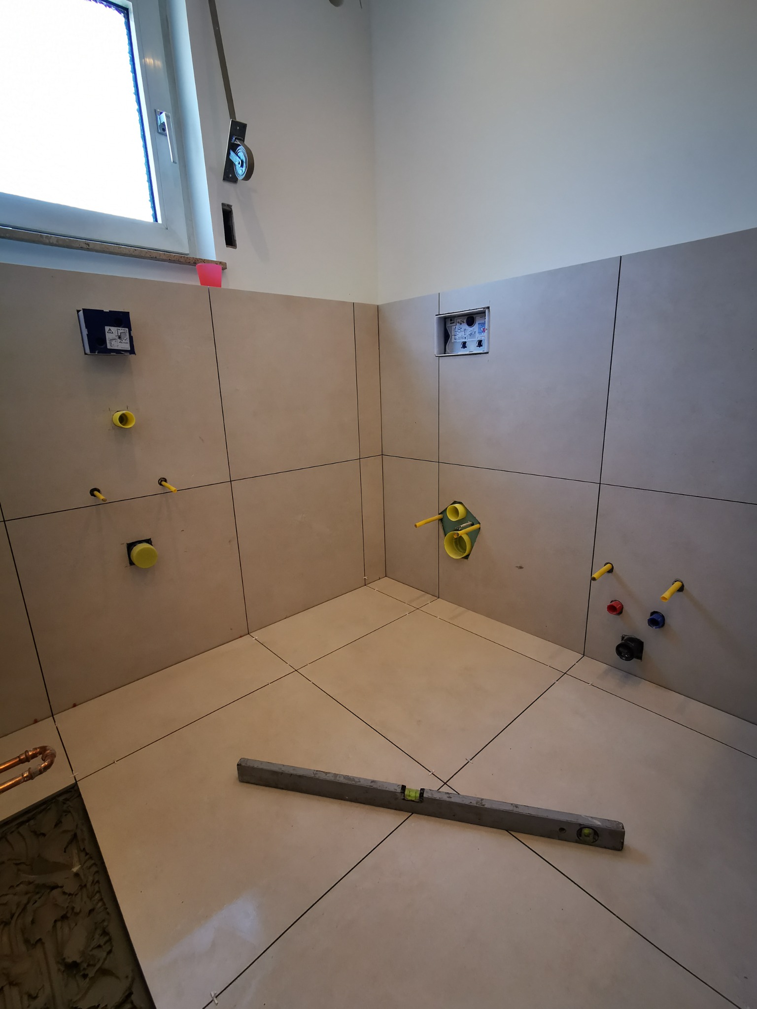 Wnętrze łazienki w trakcie układania płytek, widoczne przyłącza wodne i kanalizacyjne, poziomica na podłodze, szare płytki na ścianach i podłodze.