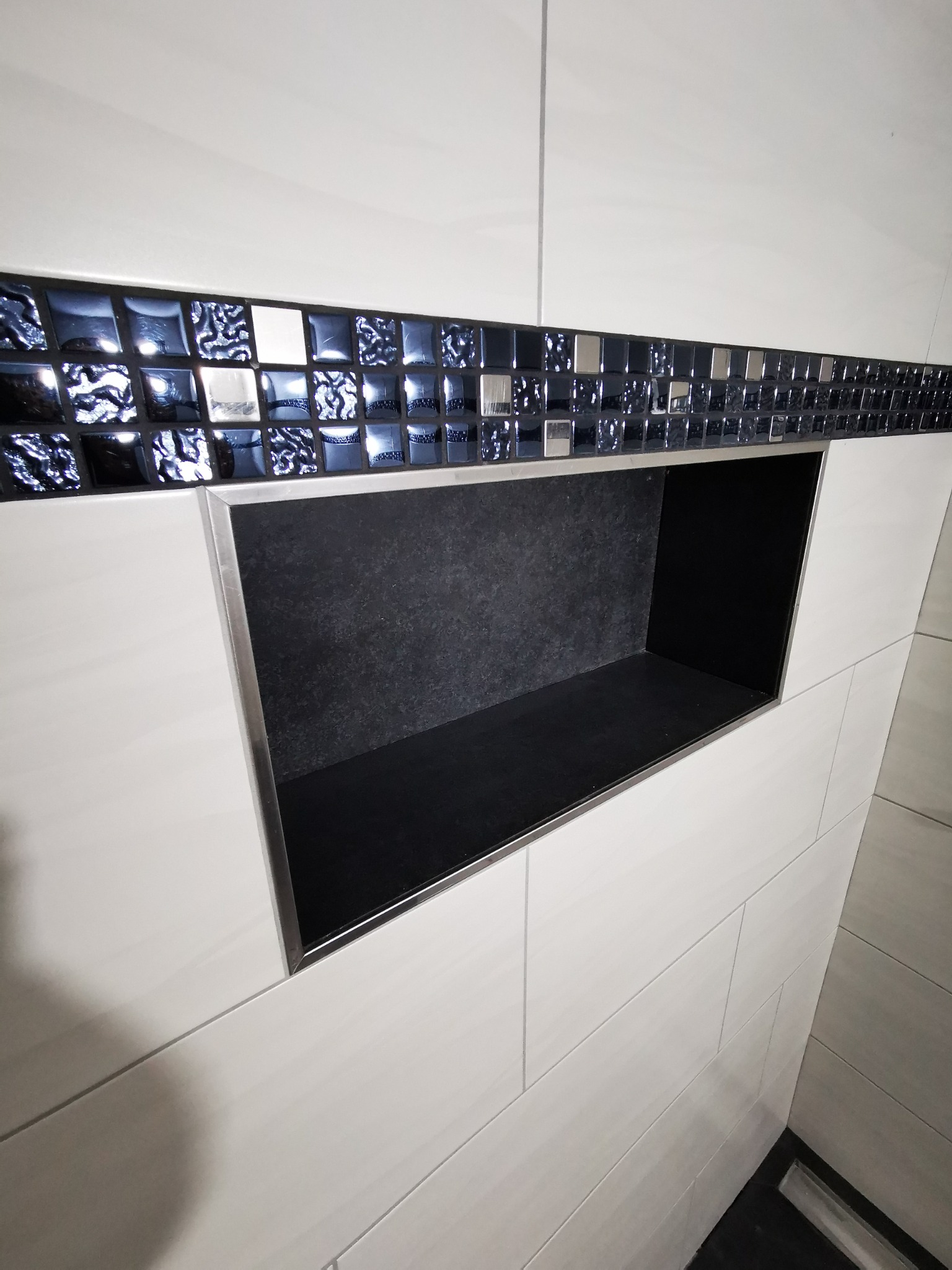 Wnęka w ścianie wyłożonej białymi, podłużnymi płytkami, obramowana mozaiką z ciemnoniebieskich i srebrnych kostek, wnętrze wnęki wykończone czarnym kamieniem.